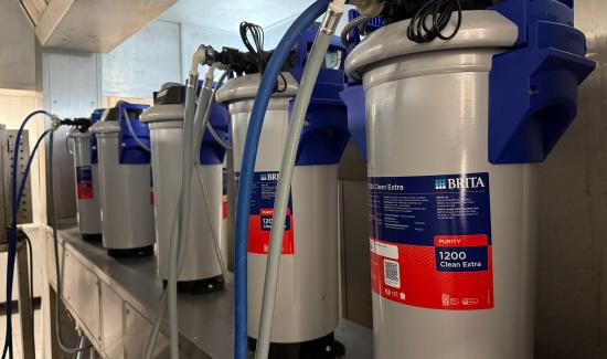 The image shows a section of a commercial kitchen installation featuring multiple BRITA Purity 1200 Clean Extra water filtration units connected by blue and grey hoses.